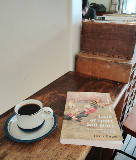 Photo is of a heavily worn wooden counter. On it is a white mug of black coffee on a white blue-rimmed saucer. The paperback book has an illustration of white rugby players in the middle of a tackle. A Japanese woman patron with bangs can be seen sitting in the distance with her face occluded.