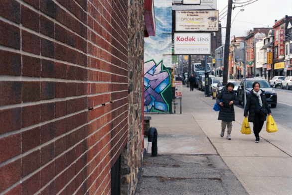 While the camera is focused on a section of street facing brick wall at an oblique angle, two pedestrians carry their groceries home along Broadview Avenue.