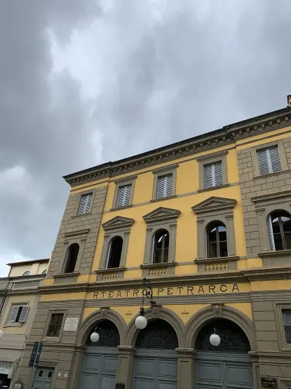 The image shows a building with a classical architectural style, featuring a yellow facade and large windows. The structure appears to be a theater, as indicated by the name "Teatro Petrarca" displayed above the entrance. The sky above is cloudy, suggesting overcast weather.
Image Credits: Wikimedia / Photo2023 / CC BY 4.0