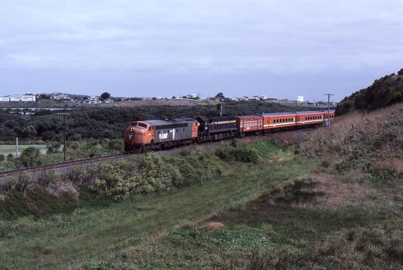 A bulldog nosed S class EMD streamlined diesel locomotive in VLine orange and grey livery and an X class hood unit in blue and gold haul a VLine passenger train in orange livery through coastal vegetation on a partly cloudy day.
