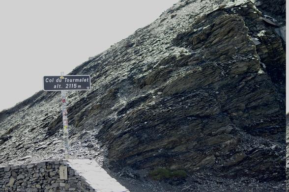 A sign indicating "Col du Tourmalet" at an altitude of 2115 meters stands against a rugged, rocky mountain terrain. The area is characterized by steep slopes and stone formations.