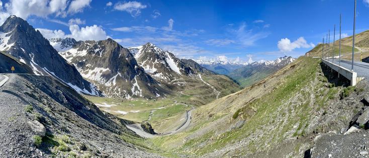 A panoramic view of a mountainous landscape featuring steep rocky slopes, patches of snow, and a winding road. The sky is blue with scattered clouds, and the scene captures the natural beauty of a high-altitude area.