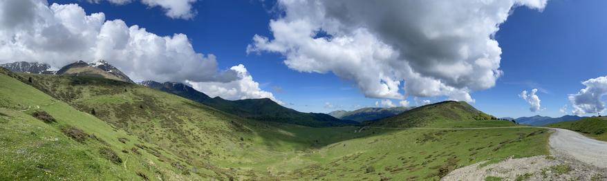 A panoramic view of a lush green landscape with rolling hills and distant mountains under a blue sky dotted with fluffy white clouds. A winding gravel path is visible in the foreground.
