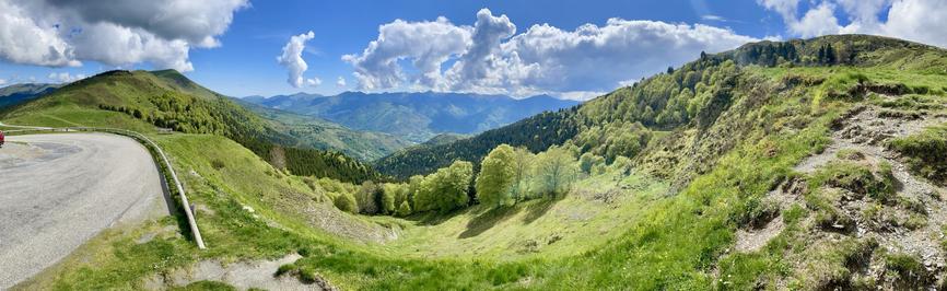 A panoramic view of green hills and valleys under a bright blue sky with fluffy clouds. A winding road is visible on the left, and a red car is parked nearby. Lush trees and mountains create a scenic natural landscape.