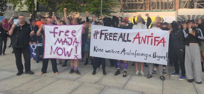 Eine Gruppe von Demonstrantinnen und Demonstranten steht auf einem Boden grauer Betonplatten. Die rechen die Fäuste und halten zwei Plakate hoch. Auf dem einen steht: "free Maja Now" auf dem anderen "Free all Antifa- keine weitere Auslieferung nach Ungarn"