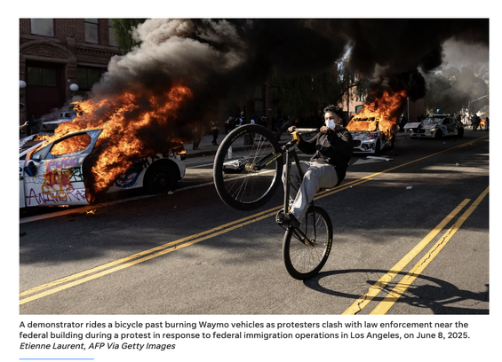 Well-composed photo of graffiti'd cars on fire. A masked person pops a wheelie on an upright bike in the double-yellow line median between those cars and the camera. The caption says "A demonstrator rides a bicycle past burning Waymo vehicles as protesters clash with law enforcement near the federal building during a protest in response to federal immigration operations in Los Angeles, on June 8, 2025.
Etienne Laurent, AFP Via Getty Images"