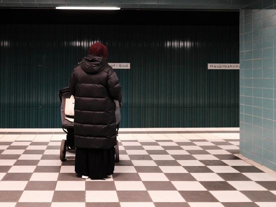 Das Bild zeigt eine Person, die mit einem Kinderwagen auf einem braun-beigefarbenen Schachbrettmusterboden in einer U-Bahn-Station steht. Die Person steht mit dem Rücken zum Betrachter. Sie trägt einen braunen, gesteppten Wintermantel, einen dunklen langen Rock und eine weinrote Mütze. Sie steht mit einem Kinderwagen auf einem braun-beigefarbenen, schachbrettförmigen Boden in einer U-Bahn-Station. Der Kinderwagen steht vor hier und ist sehr breit – möglicherweise für 2 Kinder. Die Wände der U-Bahnstation bestehen aus dunkelgrünen, vertikal gemusterten Platten und mintfarbenen Fliesen. Zwei Schilder sind nur teilweise lesbar – vermutlich steht auf beiden „Hauptbahnhof Süd“.

The picture shows a person standing with a baby carriage on a brown and beige checkerboard floor in a subway station. The person is standing with their back to the viewer. She is wearing a brown, quilted winter coat, a dark long skirt and a wine-red cap. She is standing with a baby carriage on a brown and beige checkerboard floor in a subway station. The baby carriage is in front of here and is very wide - possibly for 2 children. The walls of the subway station consist of dark green, vertically patterned panels and mint-colored tiles. Two signs are only partially legible - presumably both say “Hauptbahnhof Süd”.