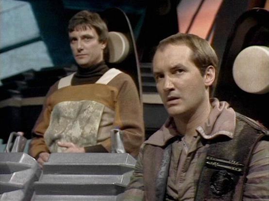 Claude Sonnet 4.0 describes the image as: "This scene appears to be set aboard a spacecraft, likely the Liberator, given the futuristic interior with curved walls and control panels visible in the background. The setting has the characteristic sleek, technological aesthetic of the series with muted lighting creating a serious atmosphere.

Two crew members are shown in what appears to be a tense or contemplative moment. One character is wearing the distinctive brown and beige Federation uniform with geometric patterns, while seated at what looks like a control station. The other character is positioned nearby, also in period-appropriate costume consisting of earth-toned clothing.

The scene captures the serious, often morally complex tone that the series was known for, with both characters appearing focused and concerned about their current situation. The spacecraft interior and their positioning suggests this is during a critical moment in their ongoing conflict with the Federation."

Blakes7Bot, version 3.9.30.