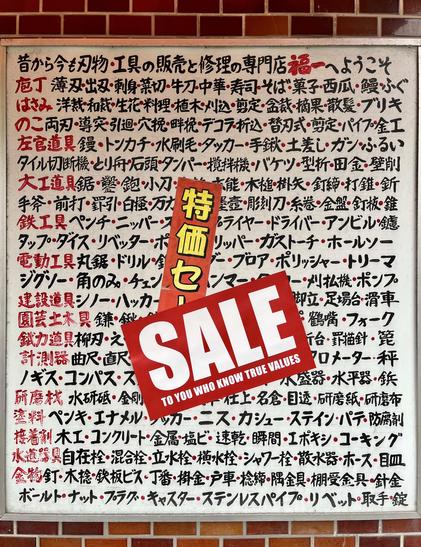 The sign of an old school Japanese knife shop, with an extremely long list of articles written by hand in black and red. On it, a first red sticker has been placed reading in yellow Japanese script“special sales”, and on top of this one, another red sticker reading in white, in English, “sale to you who know true values”.