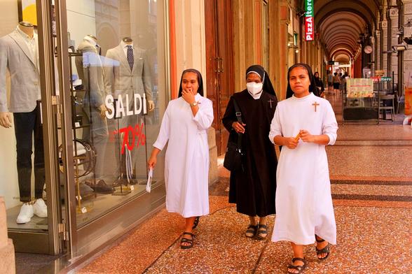 The image shows three nuns walking towards the viewer on a patterned outdoor walkway. Two are on the left and right, wearing long, white, flowing robes with short sleeves and black sandals. They have simple head coverings, and a small cross necklace is visible on the nun to the right. The third nun, in the center and slightly behind, wears a long, dark robe, also with a simple head covering, and a white face mask. She carries a dark shoulder bag. To their left is a large glass shop window displaying two male mannequins dressed in suits. A large red "SALDI" (Sales) sign with a red "70%" discount is visible on the window. In the background, a long, arched arcade stretches into the distance, lined with shops, including one with a "PizzaAltero" sign. The lighting is bright and even, giving the scene a clear, vibrant appearance.