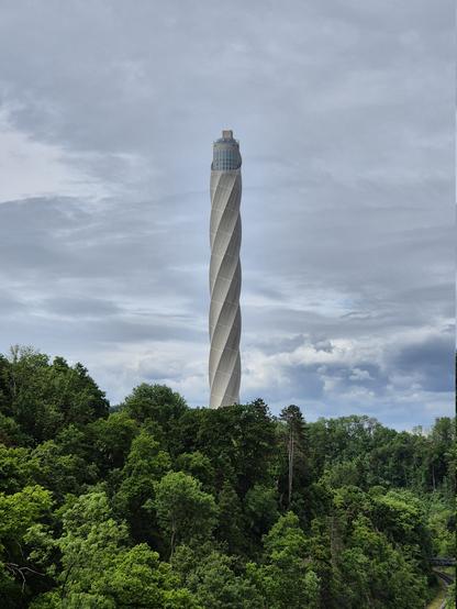 Hinter einem Wald ragt ein Turm in den bedeckten Himmel empor, der sich wie ein Schraubgewinde nach oben windet.