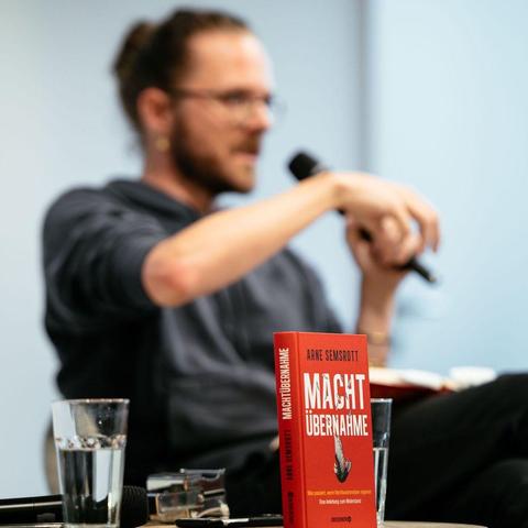 Im Fokus, das Buch von Arne Semsrott "Machtübernahme". Im Hintergrund Semsrott auf der Bühne.