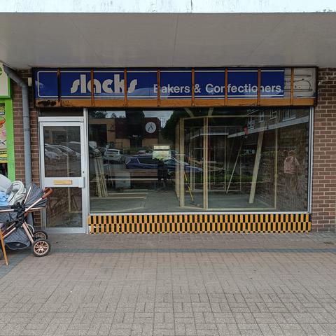 Picture of a disused shop front. Above a large glass window the framing for the shop sign has been revealed. But behind _that_ is an older sign, in white lettering on blue acrylic. "slacks / Bakers & Confectioners". "Slacks" is in one of those Bauhaus / pump / Burko fonts; "Bakers & Confectioners" is in Helvetica