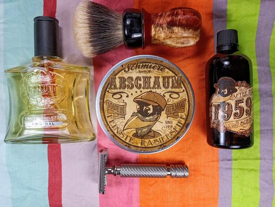 (Le Chat Mistral description)

The image showcases a collection of grooming items arranged on a striped background. At the top left, there's a bottle of Brut Original Eau de Toilette. To the right of the cologne, there's a shaving brush with a wooden handle and a shaving soap tin labelled "Abschaum" with intricate designs. Below the shaving brush, there's a safety razor with a metallic finish. On the top right, there's a bottle of aftershave with a vintage design, marked "1959." The colours are warm and inviting, with a mix of metallic and earthy tones.
