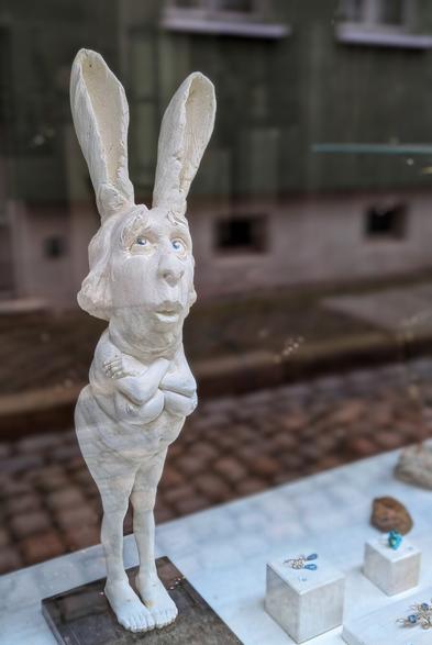 A quirky, white hare figurine with human-like features and blue eyes stands in a shop window. Its long ears are upright, and its arms are crossed over naked female breasts. The sculpture is slightly hunched, with a solemn expression. Jewellery pieces are displayed on white stands in the foreground, with a blurred street and buildings visible through the window in the background.
