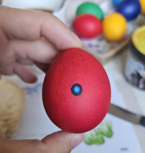 Fingers carefully holding a bright red egg. In the middle of the egg is a black circle with a bright blue spot in the center. It looks a bit like the eye of a Dalek. In the background is a table and a plate with more coloured eggs (green, yellow, blue, red).