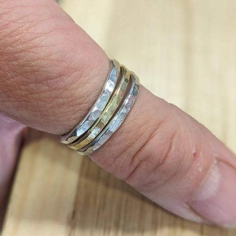 A thumb (or it might be a finger) wearing three stacking rings: two silver, one brass in the middle. The rings all have a hammered texture.