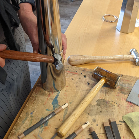A silver bangle being shaped on a mandrel using a wooden-handled hammer. A selection of other hammers and tools are scattered around, and an octagonal bangle mandrel can be seen in the background.