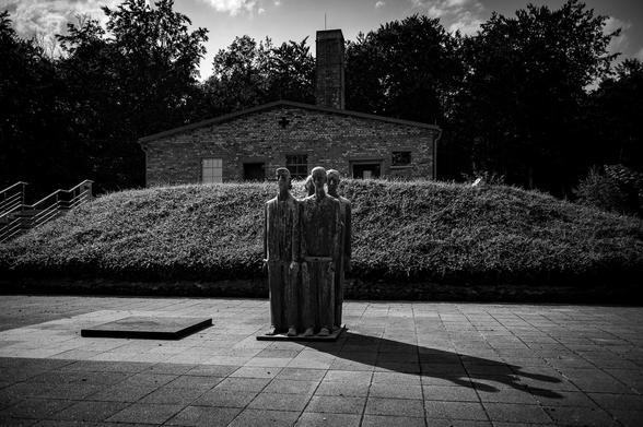 A low, stylized sculpture of several figures, presented in a static, somber pose, stands in the center of a paved area.  The figures are positioned upright, facing slightly forward, and appear to be in a group. Their forms are somewhat abstracted, and the sculptor has minimized the differences within the figures. 

A simple brick building stands in the background, positioned slightly to the rear of the sculpture. Some trees and shrubs are visible in the background.