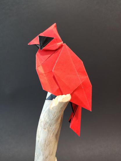 un cardinal en origami, plié dans un papier rouge et noir. Il est perché sur un support en bois fait d'une branche récupérée.