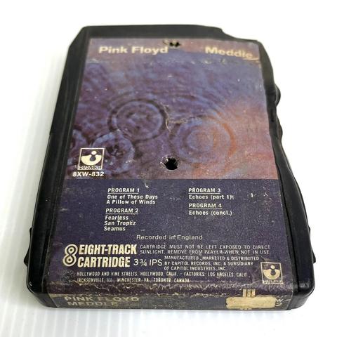 A photo of a black plastic shell 8-track cartridge tape on a white background. The tape itself is melted/deformed along its edges. The tape is entitled "Pink Floyd - Meddle" printed atop the cover art of an abstract, underwater-like image. The Harvest Records logo and track listings are toward the bottom, above a sentence that reads "EIGHT-TRACK CARTRIDGE / CARTRIDGE MUST NOT BE LEFT EXPOSED TO DIRECT SUNLIGHT. REMOVE FROM PLAYER WHEN NOT IN USE" along with Capitol Records address & copyright info.