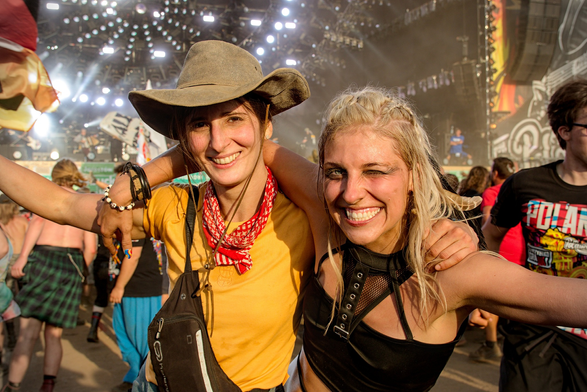 Two ecstatic festival-goers pose arm in arm, grinning from ear to ear. One wears a cowboy hat, yellow shirt, and red scarf, the other has bleached hair, smudged makeup, and a huge wink. The sun is shining, the stage is glowing, and the whole crowd behind them is alive with festival energy.