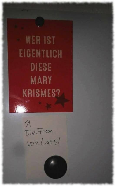 An der Wand hängt eine Postkarte mit dem Text "Wer ist eigentlich diese Mary Krismes?". Darunter hängt ein handgeschriebener Zettel mit dem Text "Die Frau von Lars!"
