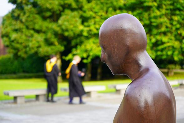 A photograph of a bronze statue of a man on a paved plaza. In the background, out of focus, are two students in black and gold graduation gowns. Beyond is a vast lawn and mature trees