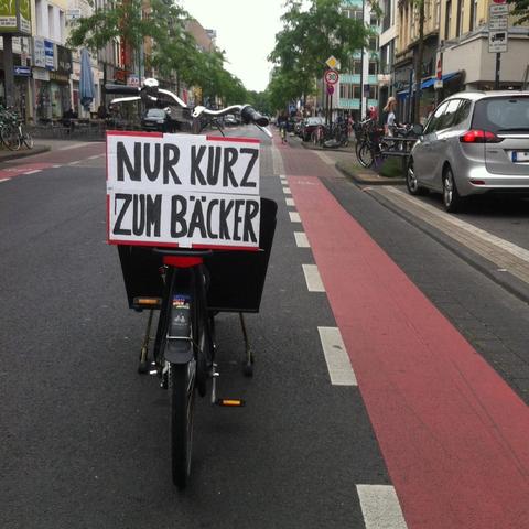 Das Bild zeigt eine Stadtstraße mit einem Fahrrad im Vordergrund, das einen großen, schwarzen Wagenanhänger trägt. Auf dem Anhänger ist ein weißes Schild mit schwarzer Schrift angebracht, das die Worte "NUR KURZ ZUM BÄCKER" trägt. Die Straße ist asphaltiert und hat eine rote Fahrradspur, die von weißen Linien markiert ist. Auf der rechten Seite der Straße sind mehrere Parkplätze mit Fahrzeugen und ein paar Fahrräder an Haltestellen zu sehen. Die Straße ist von Geschäften und Gebäuden gesäumt, und es gibt Bäume entlang der Straße. Im Hintergrund sind weitere Fahrräder und ein Auto zu sehen. Die Straße ist relativ leer, mit nur wenigen Fußgängern und Fahrzeugen.