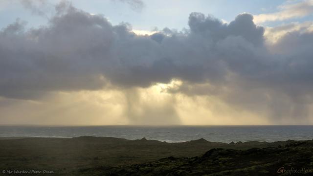 A colour photo of a landscape with the sea on the horizon. The foreground is craggy dark rocks. The sky is blue above but with a huge snow cloud in a stripe across the centre of the shot. Above it is blue sky and below is the yellowish hue of a snowstorm dropping over the ocean.