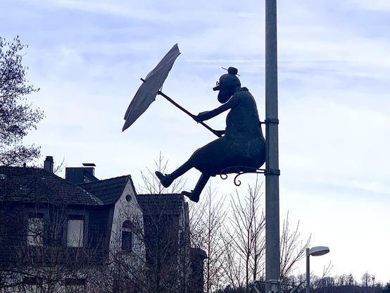 An einem Laternenmast eine Figur mit Regenschirm, direkt über der Brücke, sitzend. Februar 25 an der Lenne.
