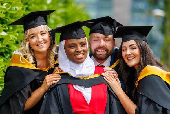 A group of graduates in black and gold academic robes and matching black mortarboard hats. They are all grouped closely together celebrating their graduation