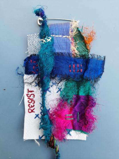 A collage art brooch, under 10 cm long and quite rectangular. On a piece of jeans fabric are applied different pieces of pure silk in blue, blueish green and some pink colours. Intuitive stitching is used to fix them. One piece is an old cross stitch embroidery in blue on white. The red word RESIST is embroidered beside the crosses. The piece is still not yet finished. All material is from reutilisation.
