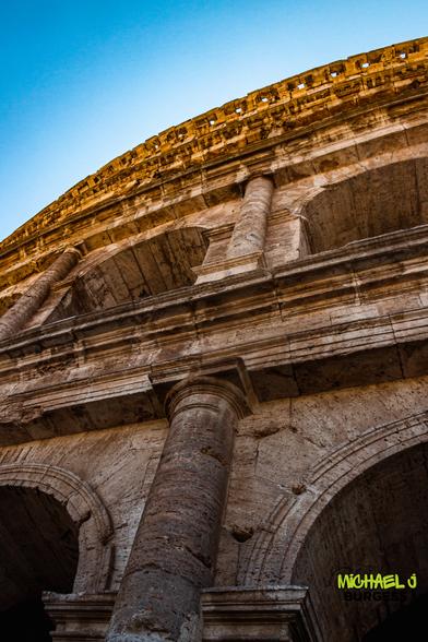 Low-angle vertical view of the Colosseum in Rome, highlighting massive stone columns and arches glowing in golden light against a clear blue sky.