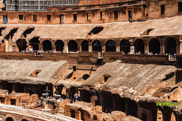 View from inside the Colosseum in Rome showing tiered seating, arched corridors lined with tourists, and remnants of ancient stonework with a cross standing on the arena floor.