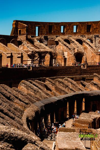 Vertical view inside Rome's Colosseum showing upper brick arches framed by blue sky, curving stone vaults and passageways below, and groups of tourists exploring the ancient amphitheatre.