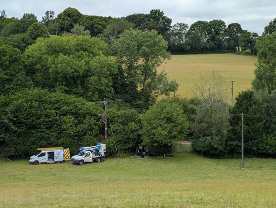 A photograph taken from a hill above a rural setting. Looking down upon two linesmen power maintenance vans. One of which has a bucket lift. The scene is very grassy and green, the trees all have green leaves.