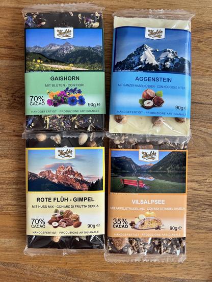 Auf dem Bild sind vier verschiedene handgefertigte Schokoladentafeln der Marke "Walde" zu sehen. Jede Tafel ist einzeln verpackt und trägt ein Etikett mit einem Foto einer alpinen Landschaft sowie dem Namen der jeweiligen Sorte. Die Sorten und ihre Besonderheiten sind:

- Oben links: "GAISHORN" – 70% Kakao, mit Blüten verfeinert.
- Oben rechts: "AGGENSTEIN" – Weiße Schokolade mit ganzen Haselnüssen.
- Unten links: "ROTE FLÜH - GIMPEL" – 70% Kakao, mit Nuss-Mix.
- Unten rechts: "VILSALPSEE" – 35% Kakao, mit Apfelstrudel-Mix.

Alle Tafeln wiegen 90 Gramm und sind als handgefertigte, handwerklich hergestellte Produkte ausgewiesen. Die Verpackungen sind farblich unterschiedlich gestaltet und zeigen jeweils eine typische Landschaft oder ein Motiv aus der Region, passend zum Namen der Sorte.