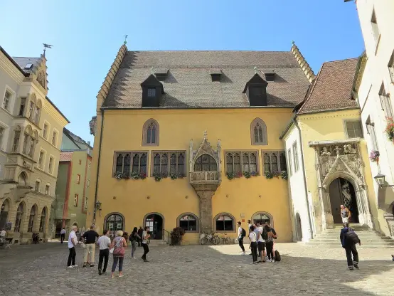 The image shows a building with a distinctive architectural style, featuring a mixture of medieval and Renaissance elements. It has a yellow facade, large windows, and decorative detailing. In front of the building, there are several people gathered, some taking photos and others walking. The setting appears to be a square or courtyard, surrounded by other structures, and the sky is clear and blue.

Image Credits: Wikimedia / Bärwinkel,Klaus / CC BY-SA 4.0