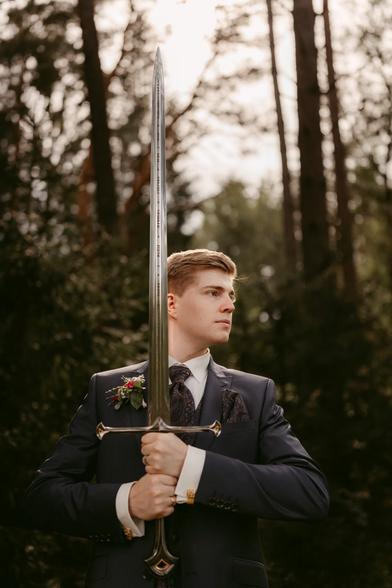 This image, taken by Hamburg based European wedding and elopement photographer Sturmsucht, captures a groom's portrait from a Lord of the Rings themed wedding. The groom is holding Andúril, the legendary reforged sword of Aragorn, vertically with both hands on the ornate hilt. The sword's distinctive design with its elaborate crossguard is clearly visible, pointing upward in a noble, ceremonial pose.  The groom is dressed in formal wedding attire - a dark charcoal suit with a white dress shirt and an elegant patterned tie or ascot. His lapel features a wedding boutonniere with red flowers and greenery. He has neatly styled short blonde hair and gazes off to the side with a serious, regal expression reminiscent of Aragorn himself.  The setting is a forest bathed in golden-hour light, creating a perfect Middle-earth atmosphere. The trees in the background are softly blurred, giving the portrait a dreamy, cinematic quality that could be straight from the films. The warm lighting creates beautiful highlights and shadows, adding to the epic fantasy feel.  This portrait beautifully combines traditional wedding formality with Lord of the Rings fandom, allowing the groom to channel his inner Aragorn on his special day. The professional photography captures both the significance of the wedding and the couple's shared love for Tolkien's world, creating a memorable image that honors both the sacred nature of marriage and their geeky passions.