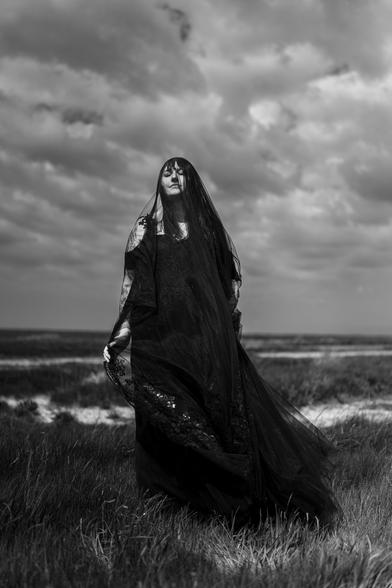This is a black-and-white photograph by Hamburg based European elopement and wedding photographer Sturmsucht showing a bride standing outdoors in a grassy field. She is wearing a long, flowing black dress and a sheer black veil that covers their head and shoulders, giving a dramatic and mysterious appearance. Her eyes are closed, and her expression is calm and serene. The sky above is filled with thick, textured clouds, adding a moody and atmospheric backdrop to the scene. The overall feeling of the image is somber and ethereal, with a strong sense of solitude and quietness.