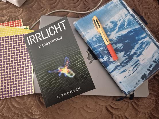 A table with some dot stickers, a copy of Irrlicht 1: Absturz by H.Thomsen and a blue hobonichi cousin book with a yellow and orange hobonichi pen.