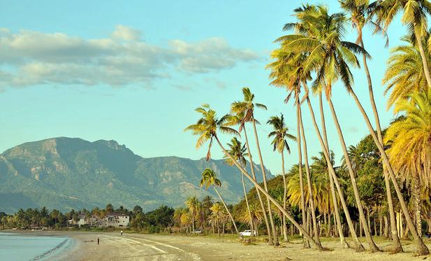 Photo shows a coarse sandy beach changing into a pebble beach before the line of palm trees breaks of the beach to the right. The beach is in a crescent towards the left with a couple of people standing and walking. The palm tree mixed with more ordinary green leafy forest stretches along the beach, and back towards a mountain range. In the forest line down to the left there is a white building, a guest house on the beach. The light is sunset warm and yellowish, and the sky is very lightly clouded light blueish indigo.