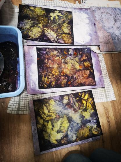 On a kitchen floor, there are some plant dyed and printed pieces of art paper. There is also a blue plastic bowl on left and the papers are on ceramic tiles on old towels.