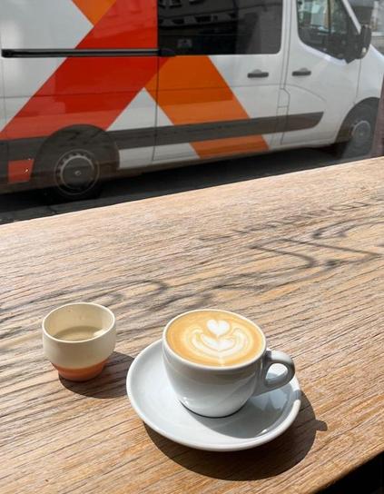 Auf einer Holzbank vor Fenster eine Tasse Cappuccino und ein getöpfertes Schälchen. Vor dem Fenster ein weiß-oranger Lieferwagen