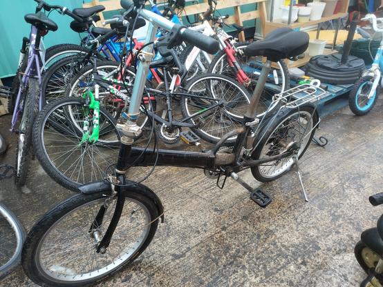 A black folding bicycle, assembled and stood up, surrounded by various other bikes in a recycling centre.