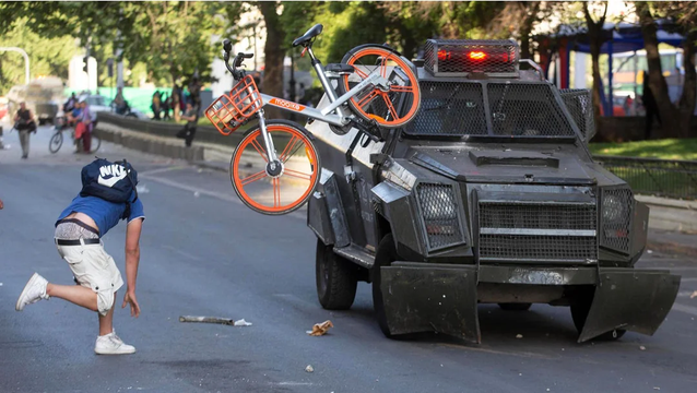 a protester with saggy clothes is throwing a bike share bike at an armored truck on a street.