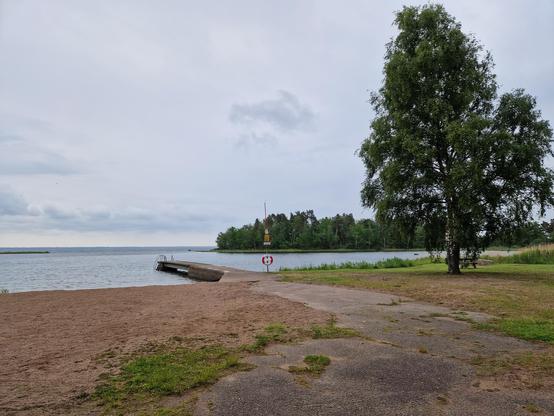 En badbrygga på lite håll. Sand och gräs på land. Bak I bild syns sjön och himmel. Ett stort träd till höger i bild.