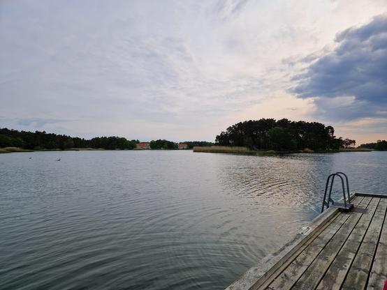 Brygga med badstege till höger i bild. Krusig vattenyta. En himmel där solen kikar fram bakom ett moln. En ö med träd bak I bilden.