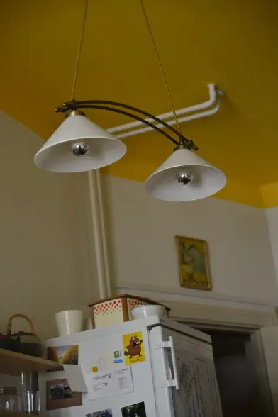 bild im hochformat in einer wohnung.
zwei rohrleitungen gehen von der decke an der wand herunter. von der decke eine lampe mit zwei lampenschrimen.
in allem zusammen könnte man karl klammer erkennen